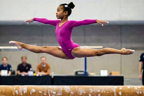 A gymnast in a split leap above the beam, with the judges' table behind her, seen from across the hall.